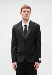 Μαύρο σακάκι tuxedo με σατέν πέτα, κλείσιμο με ένα κουμπί και δύο εμπρός τσέπες. Φοριέται πάνω από πουκάμισο με λευκές ρίγες και μαύρη γραβάτα.