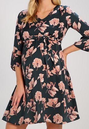 Frau trägt ein dunkelgrünes, florales Umstandskleid mit rosa Blumen, dreiviertellangen Ärmeln, V-Ausschnitt und gebundenem Taillenband.