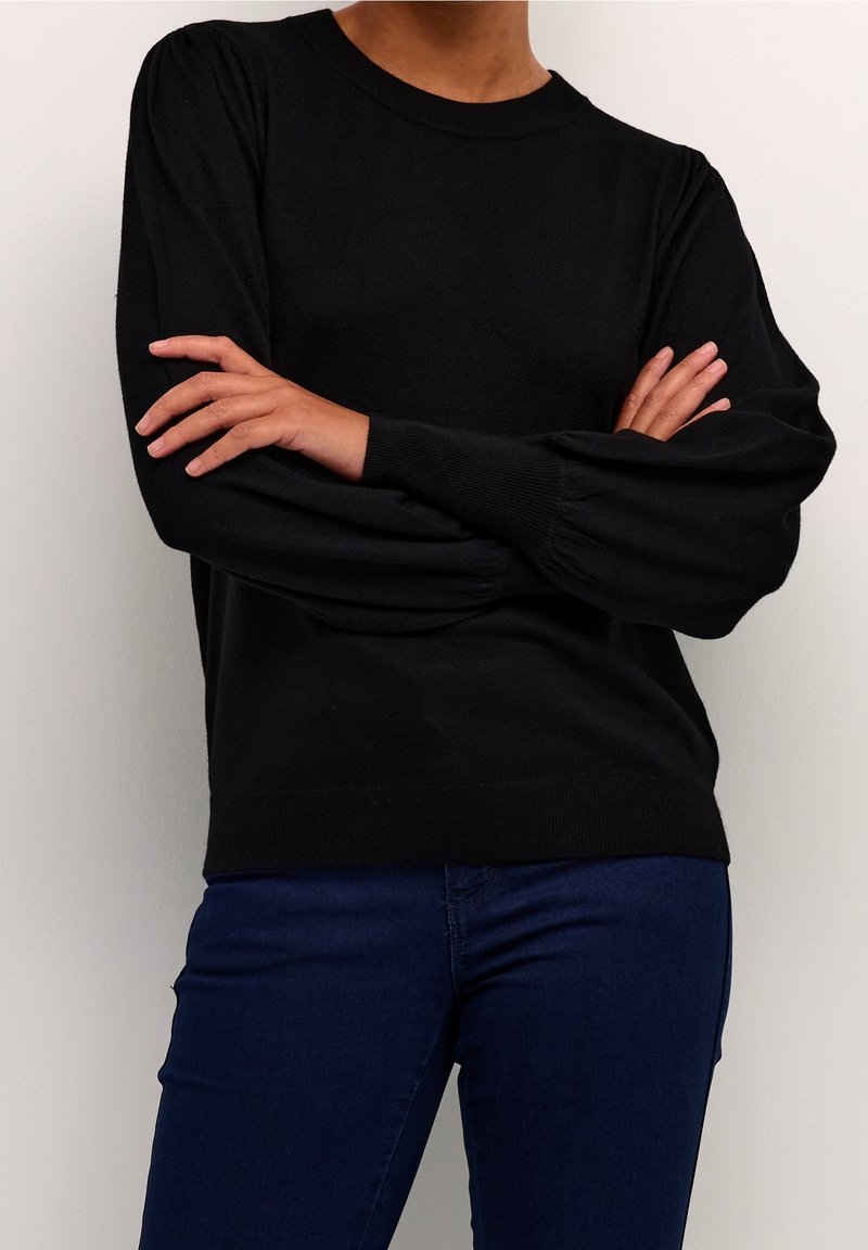 Black sweater with long, gently puffed sleeves, ribbed cuffs, and a round neckline. Worn with dark blue jeans against a neutral background.