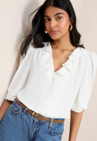 Žena s dlouhými černými vlasy, oblečená v zlatých kruhových náušnicích, bílé blouse se volánky, modrých džínách a hnědém pásku, stojící proti světlé zdi.