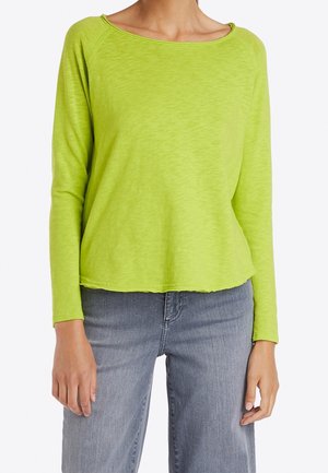 Kobieta ubrana w limonkowo-zielony, długi rękaw z szerokim, okrągłym dekoltem oraz jasnoszare dżinsy, stojąca na jednolitym tle.