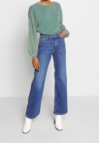 Blusa verde con estampado y mangas abullonadas, combinada con pantalones de mezclilla anchos de tiro alto en azul y botines de tacón blanco.