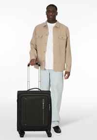 Valigia nera con superficie testurizzata, tasca frontale con zip, dettagli metallici e manico retrattile. Ruote scorrevoli.