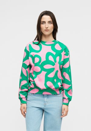 Hosszú barna hajú nő, aki rózsaszín és zöld absztrakt mintás pulóvert és világoskék farmert visel, és egy egyszerű világos háttér előtt áll.