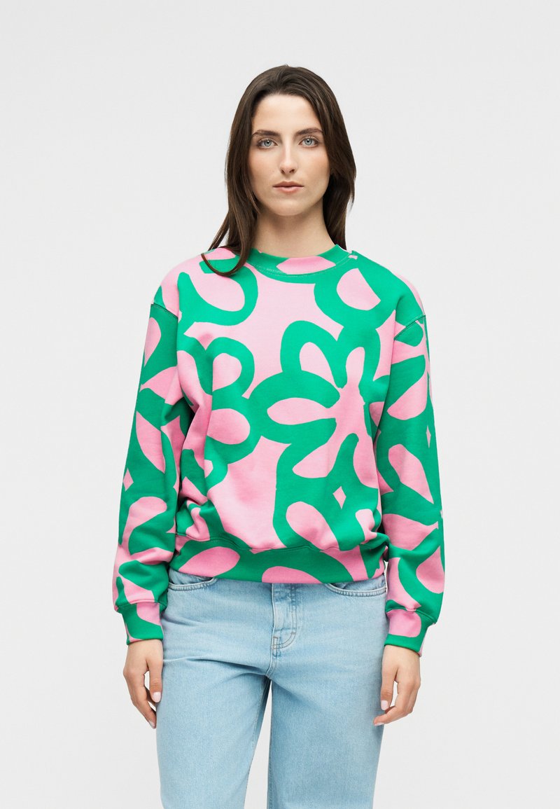 Žena s dugom smeđom kosom koja nosi majicu s apstraktnim uzorkom u ružičastim i zelenim tonovima te svijetloplave traperice, stoji na neutralnoj svijetloj pozadini.