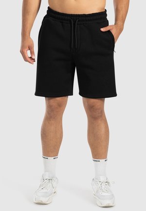 Pantalones cortos negros de algodón con cintura elástica y cordón, con bolsillos laterales y un corte relajado. Combinados con zapatillas blancas y calcetines.