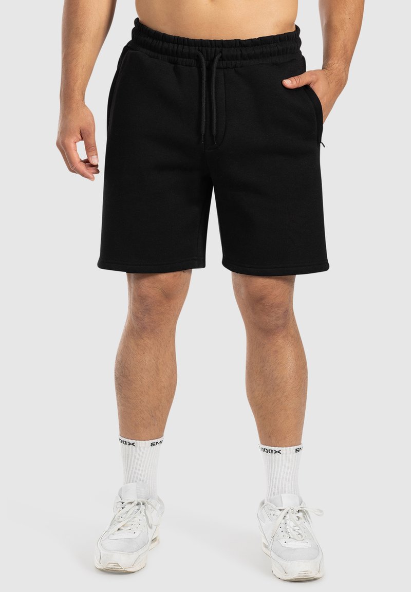 Pantalones cortos negros de algodón con cintura elástica y cordón, con bolsillos laterales y un corte relajado. Combinados con zapatillas blancas y calcetines.