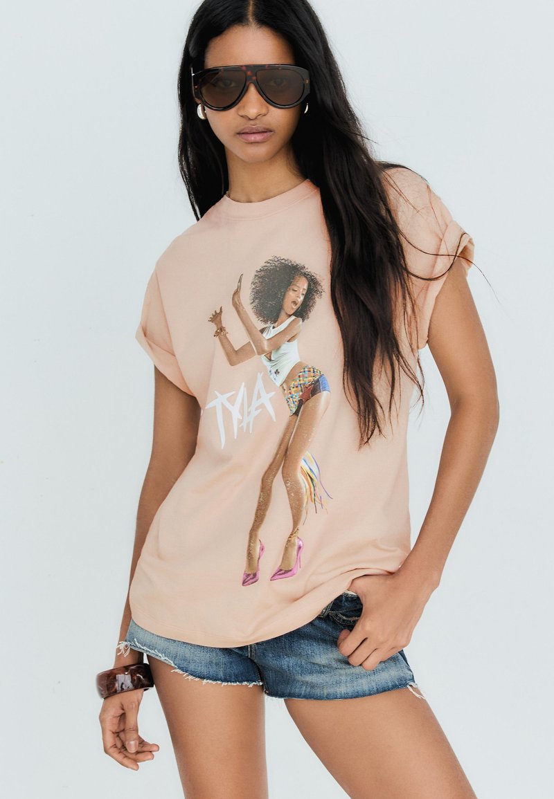 Jeune femme aux longs cheveux foncés portant des lunettes de soleil oversize, un t-shirt graphique pêche avec un motif de femme dansante, un short en jean et un bracelet.