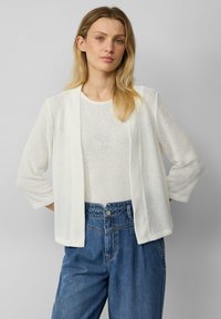 Weißer gestrickter Cardigan mit offenem Vorderteil und dreiviertellangen Ärmeln, getragen über einem strukturierten weißen Top und hochgeschnittenen blauen Jeans.