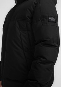 Giacca imbottita nera con cappuccio, dotata di una etichetta patch sul braccio sinistro, tasche laterali e una texture liscia e opaca. Il design è minimalista.