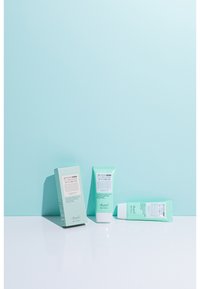 Benton UV Defense Solkräm i grön tub, bredvid förpackad produkt med vit och rosa text. Slät konsistens på en vit yta mot en blå bakgrund.