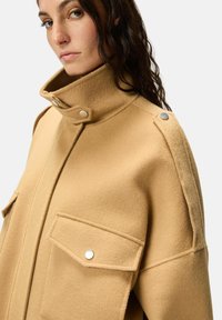 Manteau en mélange de laine beige avec col montant, fermetures à pression et deux poches avant. Finition texturée avec des accents gris contrastants.