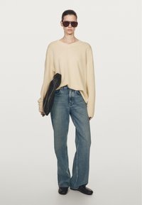 Vrouw met beige oversized trui, blauwe rechte jeans, zwarte schoenen, grote zonnebril, en een zwarte clutch in haar hand tegen een effen achtergrond.