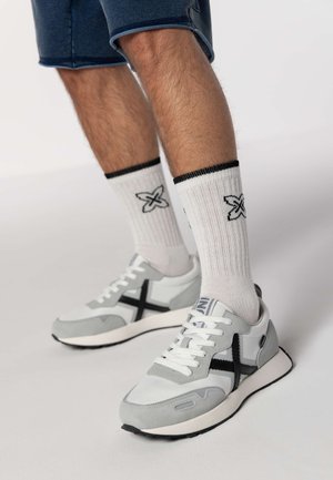 Sneaker grigie e bianche con accenti neri, caratterizzate da una tomaia in mesh, suede testurizzato e lacci bianchi. Indossate con calzini bianchi a coste.