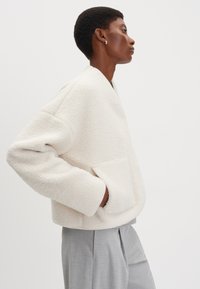 Giacca in pile color crema con vestibilità comoda, maniche larghe e tasca frontale. Abbinata a pantaloni grigio chiaro. Tessuto dalla texture liscia.