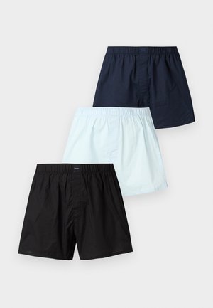 Mężczyzna bez koszulki w granatowych szortach stoi boso z jedną ręką na biodrze, obok ciemnozielonego skórzanego fotela na tle gładkiej białej ściany.