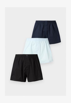 Ing nélkül álló férfi tengerészkék rövidnadrágban, egyik kezét a csípőjére téve, mezítláb, mellette egy sötétzöld bőr fotel, mögötte sima fehér fal.