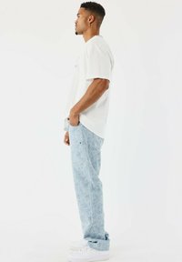 Joven de pie de perfil, vistiendo una camiseta blanca, jeans de patrón azul claro y zapatillas blancas frente a un fondo blanco liso.