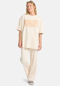 Creme oversized t-shirt med orange blomster grafik, parret med strukturerede cremefarvet brede bukser. Casual, afslappet pasform og blødt stof.