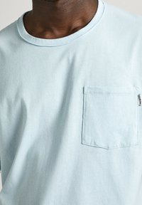 Lyseblå bomulds t-shirt med rund hals, der har en enkelt påsyet lomme på venstre side, glat stoftekstur og syet detaljer.