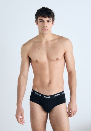Schwarze Baumwollslips mit einem weißen "HUGO"-Logo am Bund, die einen engen Sitz und eine glatte Textur bieten. Das Model steht frontal.