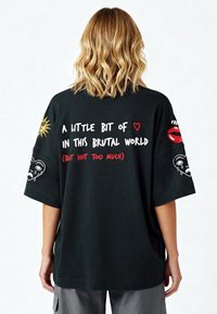 Melna pārmērīga t-kreklu ar krāsainiem izšūtiem uzrakstiem un dizainiem, tostarp saule, sirdis un sejas uz aizmugures un piedurknēm.