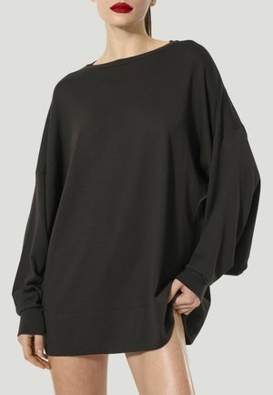 Donna con indosso un top nero a maniche lunghe dal taglio ampio con scollo a barca, che tiene l'orlo, mostrando gambe nude e rossetto rosso su sfondo neutro.