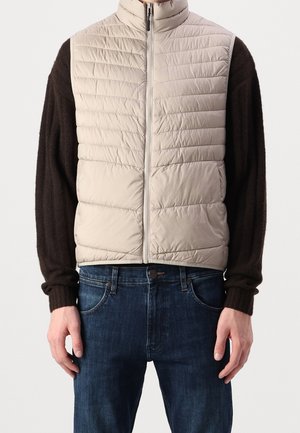 Veste beige matelassée sans manches avec fermeture éclair à l'avant portée sur un pull noir à manches longues et un jean bleu, montrée de la taille au cou.