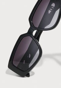 Gafas de sol negras en forma de cuadrado con lentes oscuros, con montura gruesa y un acabado brillante. Diseñadas con una forma geométrica audaz.