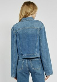 Krótka kurtka dżinsowa w jasnoniebieskim kolorze, z klasycznym kołnierzykiem, długimi rękawami i przeszyciem na plecach. Materiał ma miękką, teksturowaną powierzchnię.