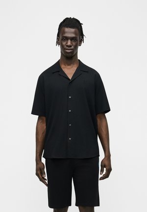 Mand med korte dreadlocks iført en sort kortærmet skjorte med knapper og sorte shorts, stående mod en ensfarvet lys baggrund.