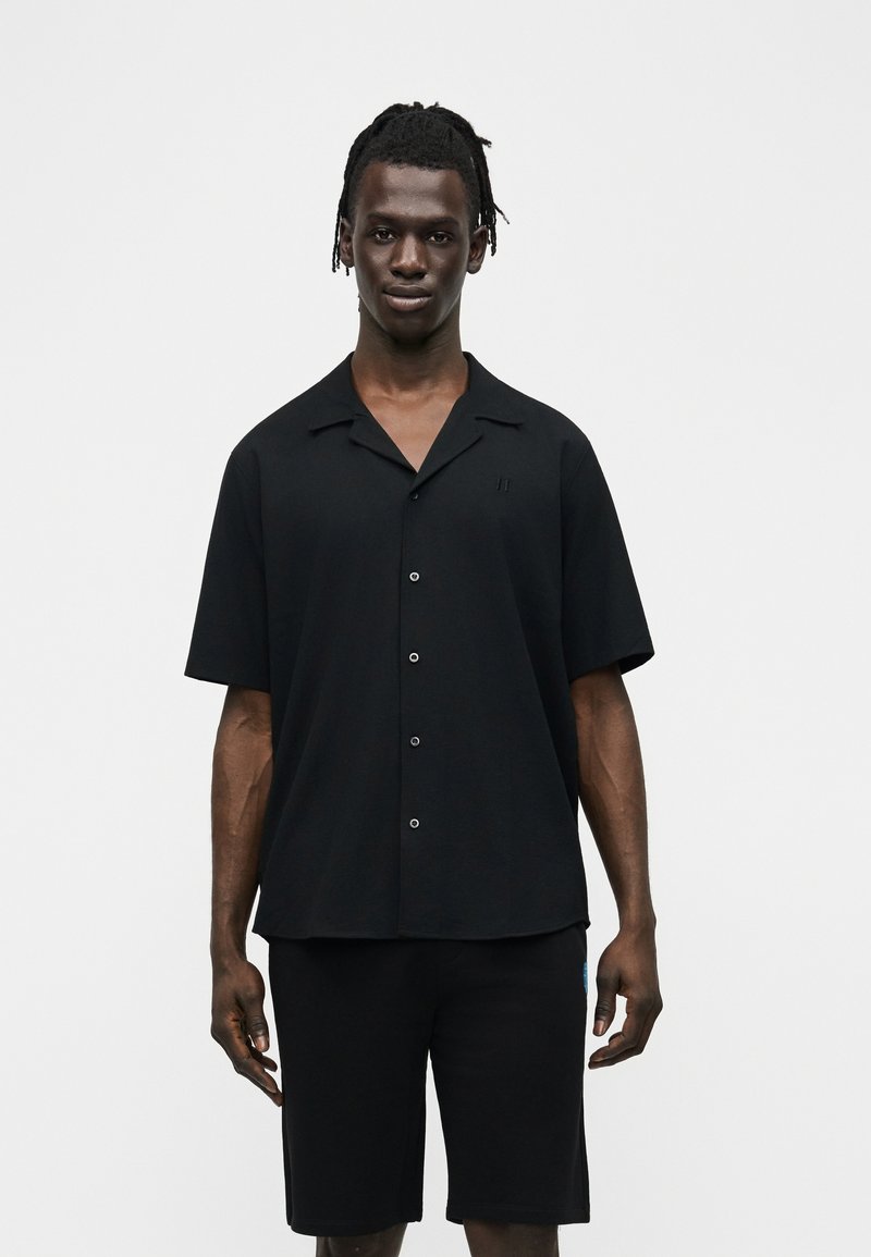 Άνδρας με κοντά ντρέντλοκς, ντυμένος με μαύρο πουκάμισο με κοντά μανίκια και μαύρο σορτς, στέκεται μπροστά σε απλό ανοιχτόχρωμο φόντο.