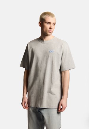 Młody mężczyzna z krótkimi blond włosami, ubraną jasnoszarą oversize koszulkę z małym niebieskim logo nieskończoności oraz jasnoniebieskie dżinsy, patrzący w prawo.