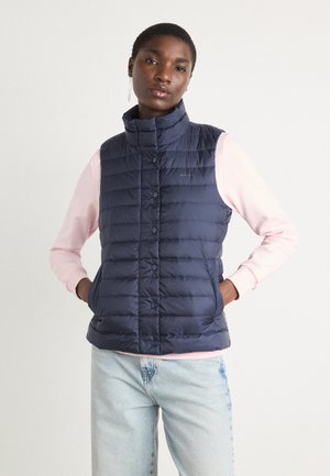 Marineblaue Steppweste mit hohem Kragen, mit gestepptem Material und Druckknopf-Verschlüssen, getragen über einem hellrosa Langarmshirt.