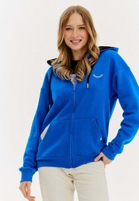 Niebieska bluza z zamkiem błyskawicznym, z kontrastującym czarnym kapturem, kieszeniami z przodu i białym graficznym detalem w kształcie rzęs. Wykonana z miękkiej, teksturowanej tkaniny.