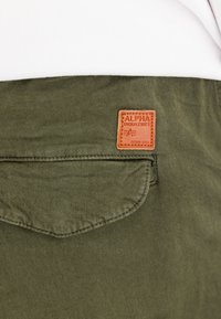 Olijfgroene broek met een bruine leren label met de tekst "ALPHA INDUSTRIES". De stof heeft een gladde structuur met een detail van een achterzak.