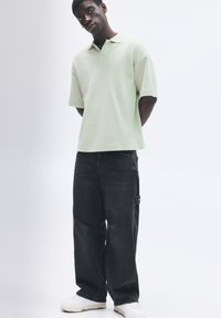 Lys grøn polo-shirt med løs pasform, korte ærmer og krave; parret med mørke, baggy jeans og hvide sneakers.