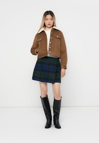 Bruine suède cropped jack met een witte schapenvachtkraag, groen-blauw geruite rok en zwarte kniehoge laarzen.