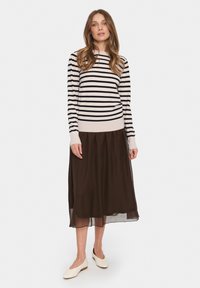 Gestreifter Pullover in Beige und Schwarz, kombiniert mit einem dunkelbraunen Faltenrock. Glatte Textur und dezente Schichtung, ergänzt durch beigen Ballerinas.