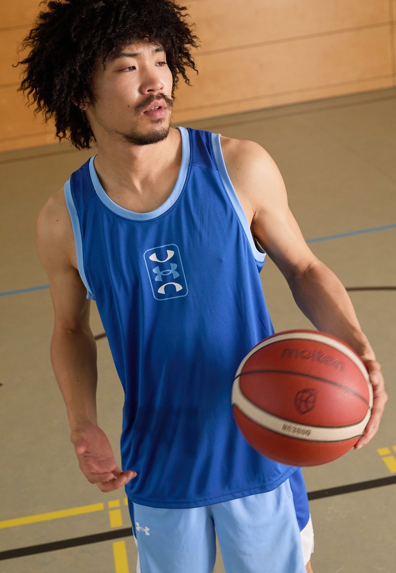 Niebieski top sportowy z jasnoniebieskimi akcentami, z logo z przodu. Model trzyma czerwoną i białą piłkę koszykową. Indoorowe ustawienie sportowe.