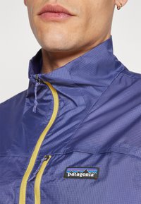 Giacca Patagonia blu navy con colletto alto, zip gialla e tessuto texture. Presenta un logo sul petto e una tasca frontale con zip.