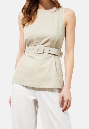 Beżowa bluzka bez rękawów z szerokim pasem w tym samym kolorze, noszona z białymi szerokimi spodniami, na modelce na jednolitym tle.