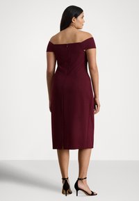 Μπορντό φόρεμα με έναν ώμο, εφαρμοστή γραμμή, μήκος μέχρι το γόνατο και φερμουάρ στο πίσω μέρος· συνδυασμένο με μαύρες γόβες με λουράκι στον αστράγαλο.