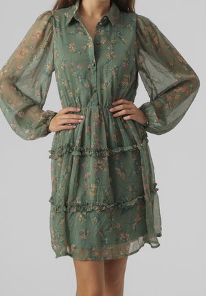 Femme portant une robe verte transparente à fleurs avec des manches longues bouffantes, un col, une fermeture boutonnée à l'avant et une jupe à volants en plusieurs couches, mains sur les hanches.