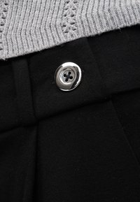 Ασημένιο κουμπί που κλείνει μαύρο ύφασμα κάτω από ένα ανοιχτό γκρι πλεκτό ένδυμα με κάθετο και δαντελωτό μοτίβο πλέξης.