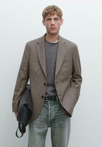 Brown checked blazer with notch lapels, two buttons, and side pockets; paired with a grey sweater and faded blue jeans. Black bag on arm.