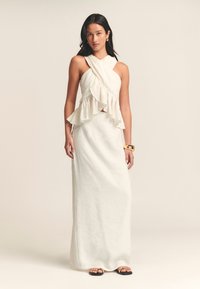 Robe maxi blanche sans manches avec un ourlet asymétrique à volants et un devant croisé. Tissu lisse avec une texture légère. Sandales noires avec des accents dorés.