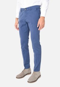 Pantaloni di cotone blu con una vestibilità slim e davanti piatto, dotati di tasche frontali e chiusura a un bottone. Indossati con stivaletti beige alla caviglia.