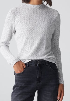Personne portant une chemise à manches longues texturée gris clair et un jean sombre, avec une main dans la poche, sur un fond neutre.