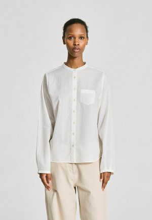 Personne debout face à l’avant, portant une chemise blanche à boutons avec une poche poitrine et un pantalon beige, sur un fond uni clair.